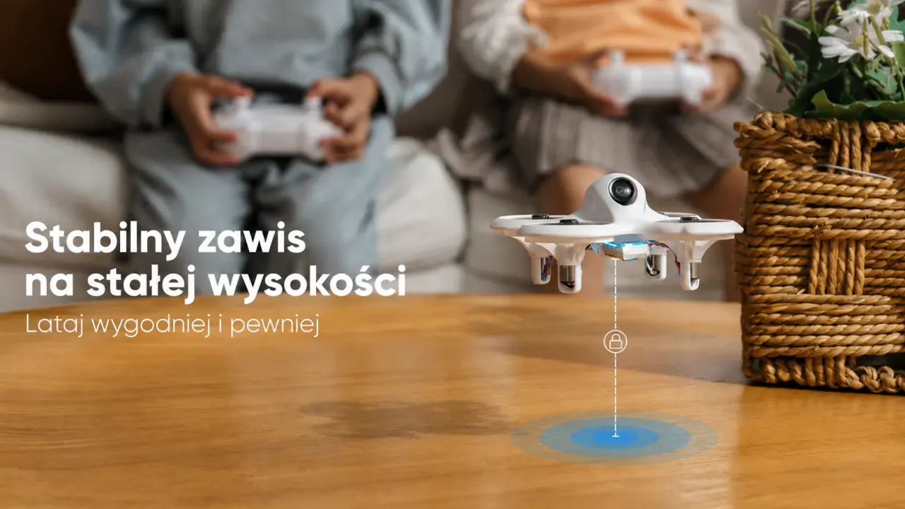 Biały mini dron FPV w zawisie nad stołem, w tle osoby trzymające kontrolery.