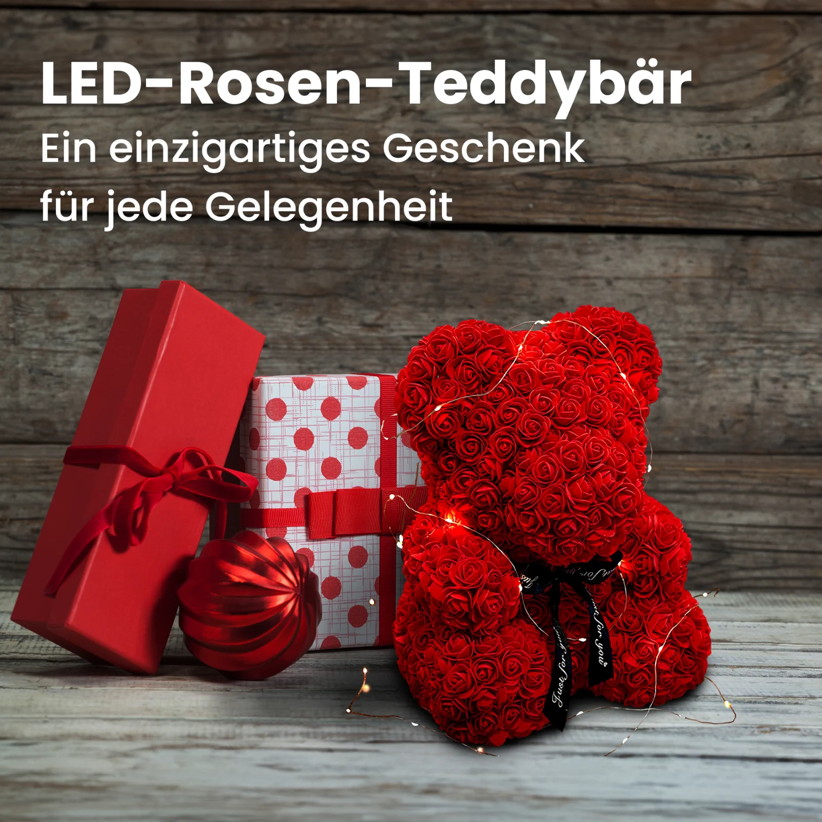 Ein roter Rosen-Teddybär, eingewickelt in LED-Lichter, steht neben zwei Geschenkboxen und einer Weihnachtskugel