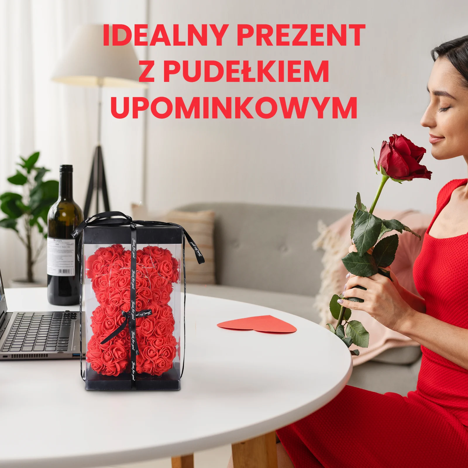 Una mujer con un vestido rojo huele una rosa roja, sentada a una mesa con un osito de peluche hecho de rosas, envuelto en una caja transparente