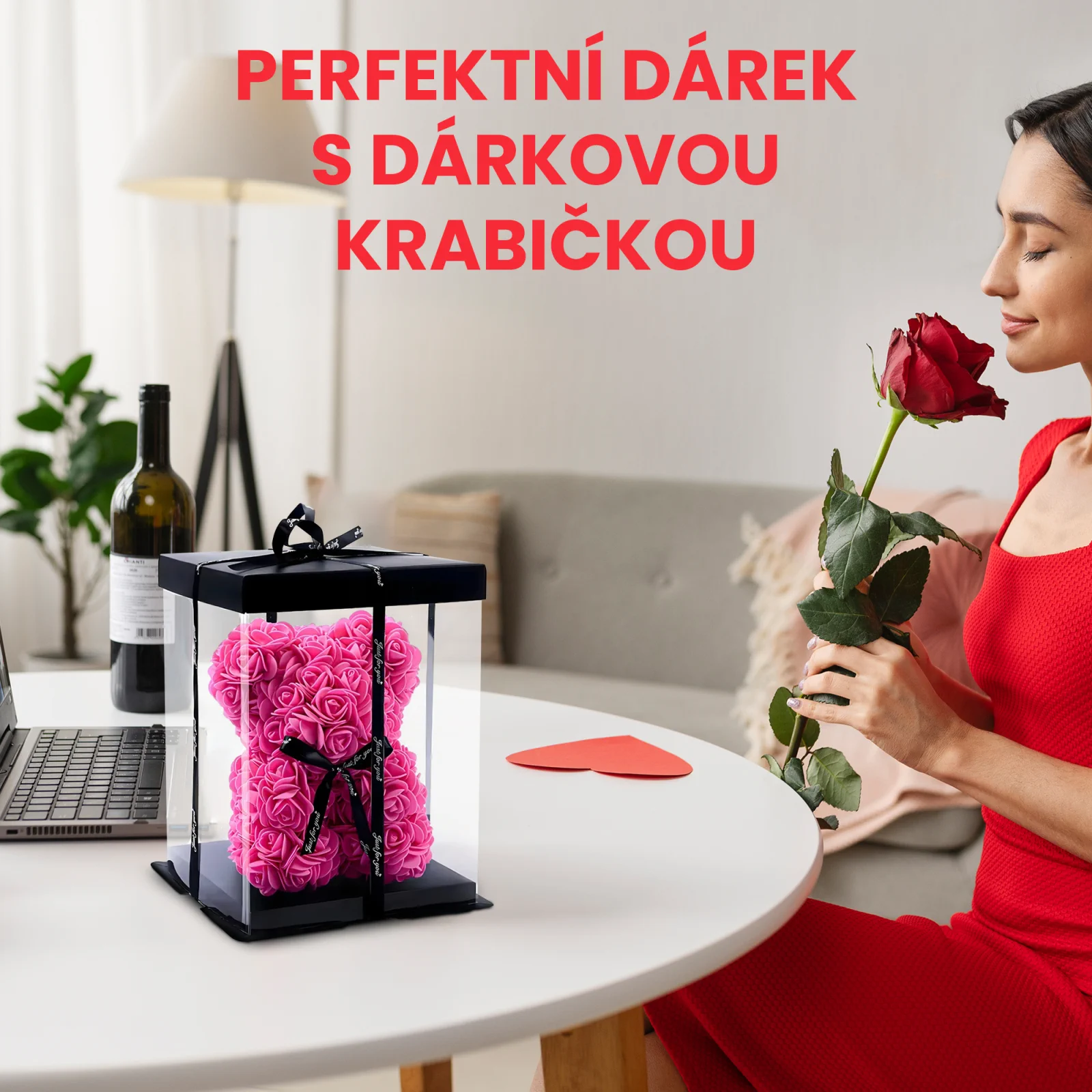 Žena v šatech čichá k červené růži a sedí u stolu s medvídkem z růží zabaleným v průhledné krabici.