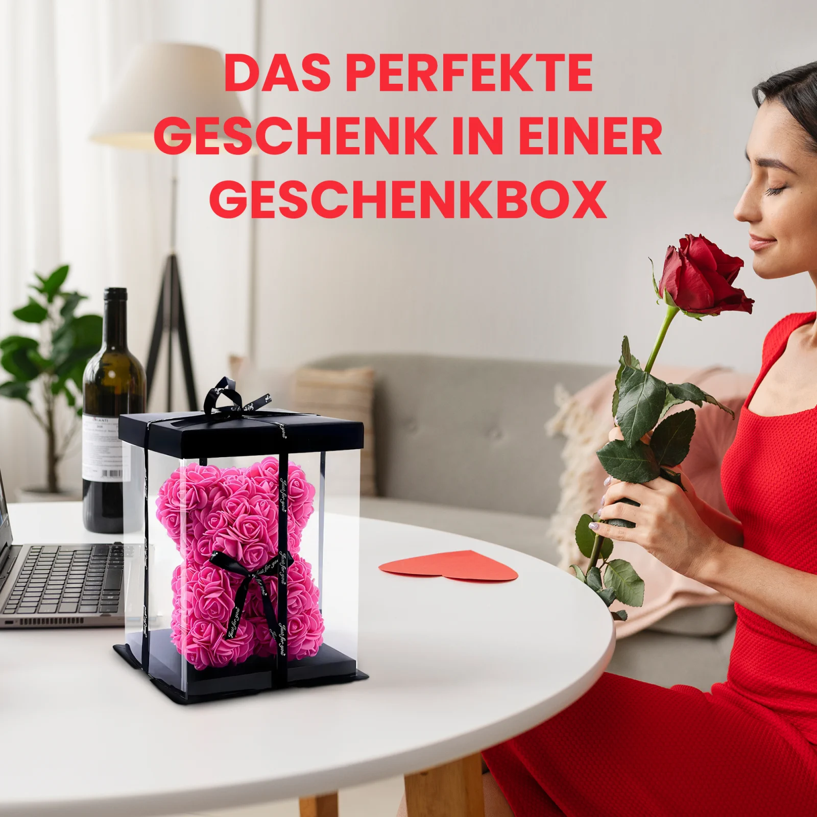Eine Frau im Kleid schnuppert an einer roten Rose, während sie mit einem Teddybären aus Rosen, der in einer durchsichtigen Schachtel verpackt ist, am Tisch sitzt