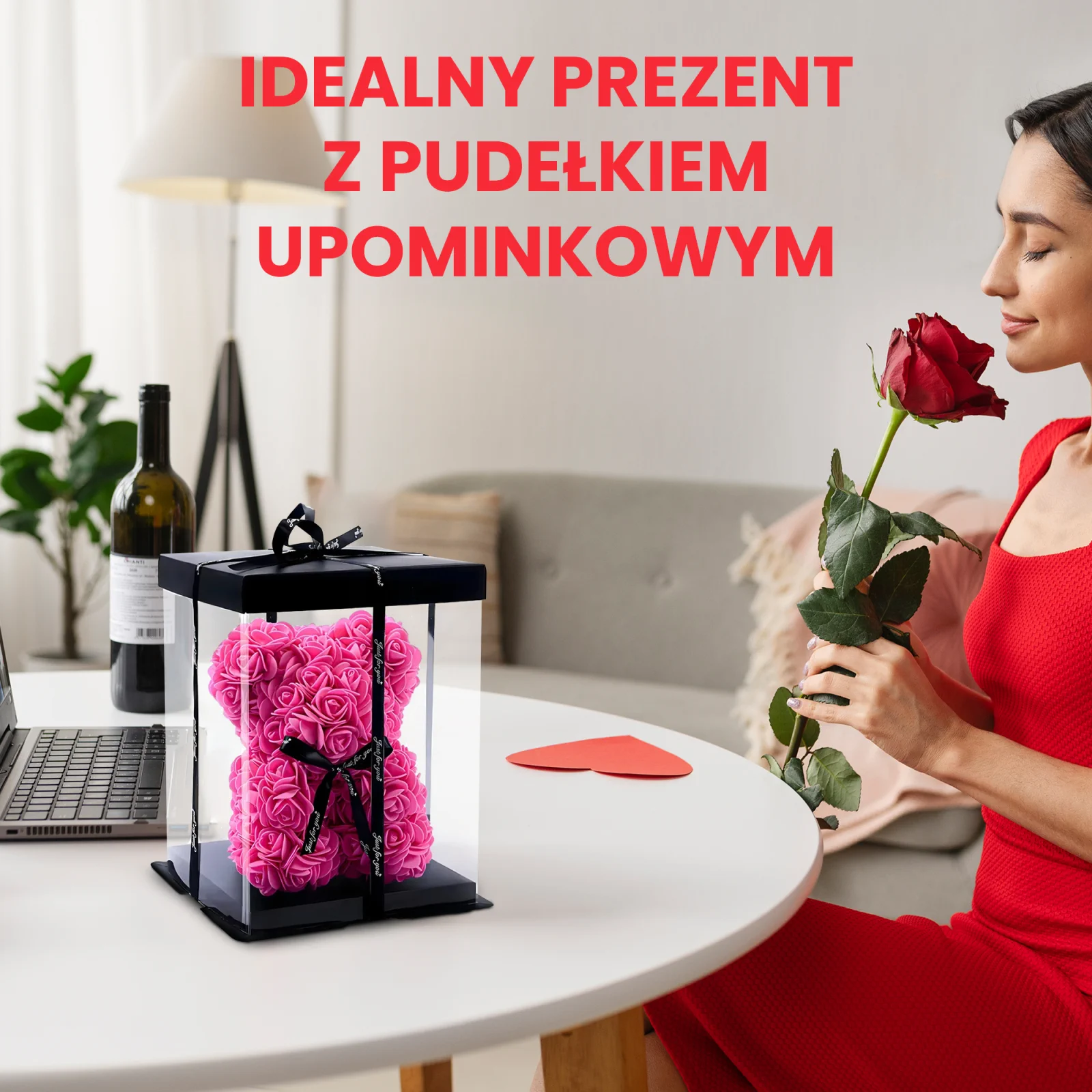 Kobieta w sukience wącha czerwoną różę, siedząc przy stole z misiem z róż, zapakowanym w przezroczyste pudełko