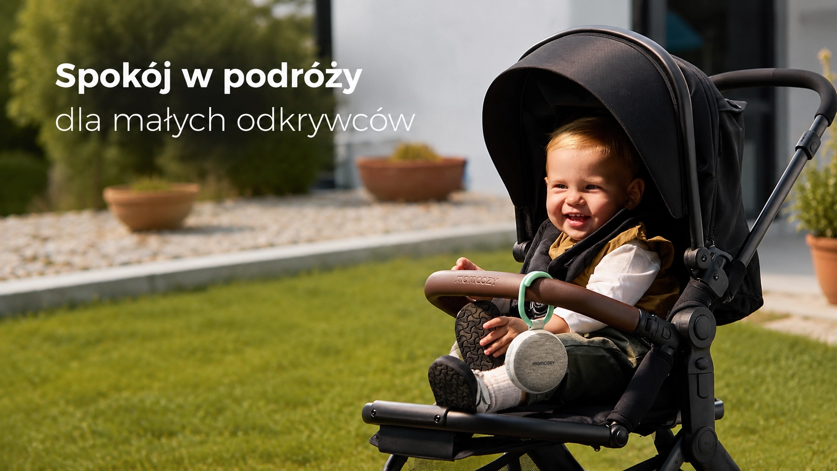 Улыбающийся ребенок в коляске с устройством Momcozy, прикрепленным к дуге. Солнечный день в саду.
