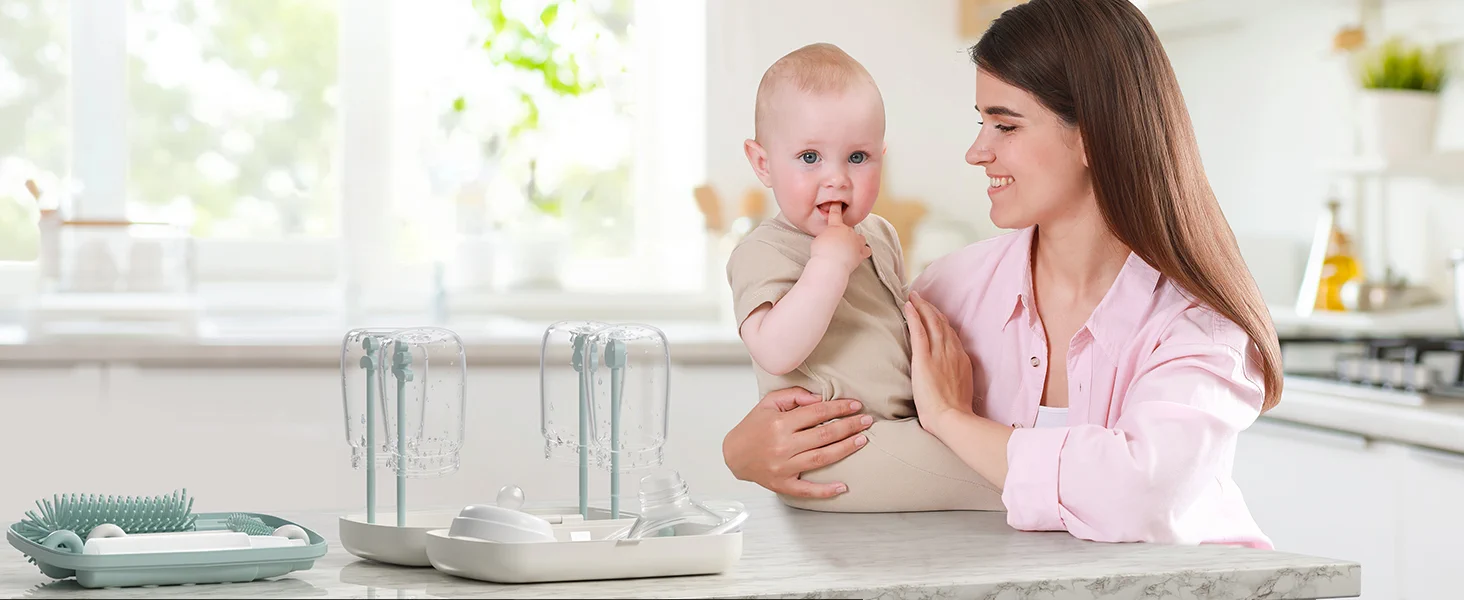 Mulher sorridente segurando um bebé na cozinha ao lado de um conjunto de lavagem e secagem de biberões montado