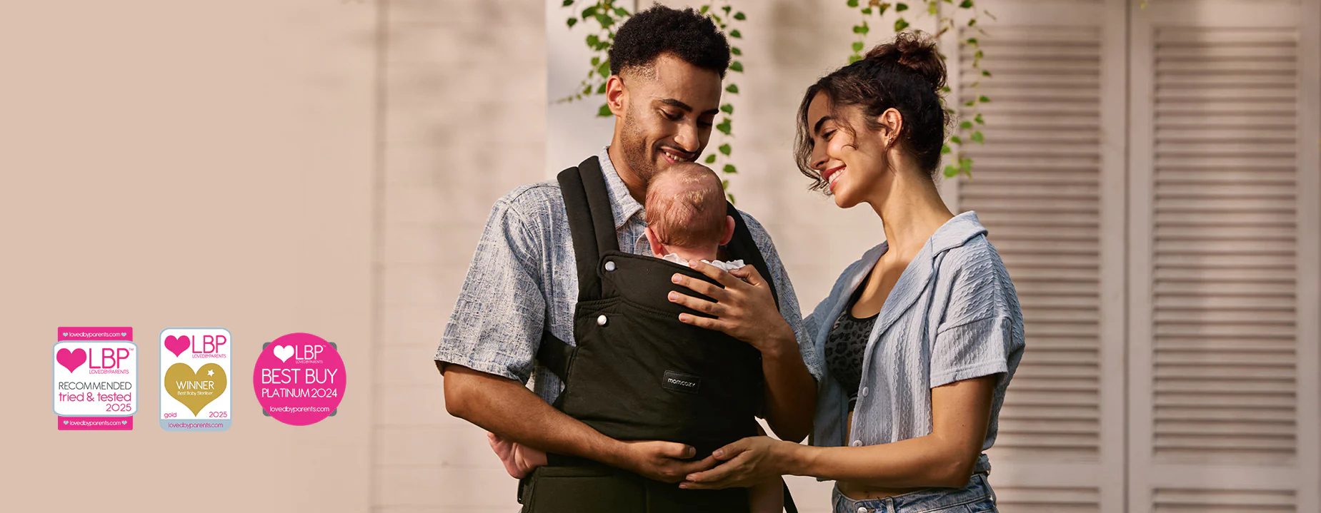 Homem e mulher sorriem para um bebê em um porta-bebês preto, com uma parede clara ao fundo.