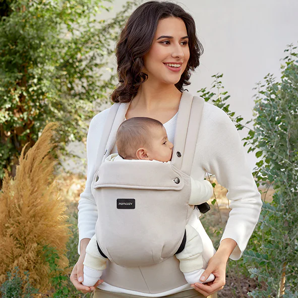 Una donna in un maglione bianco porta un neonato in un marsupio beige sullo sfondo di cespugli verdi.