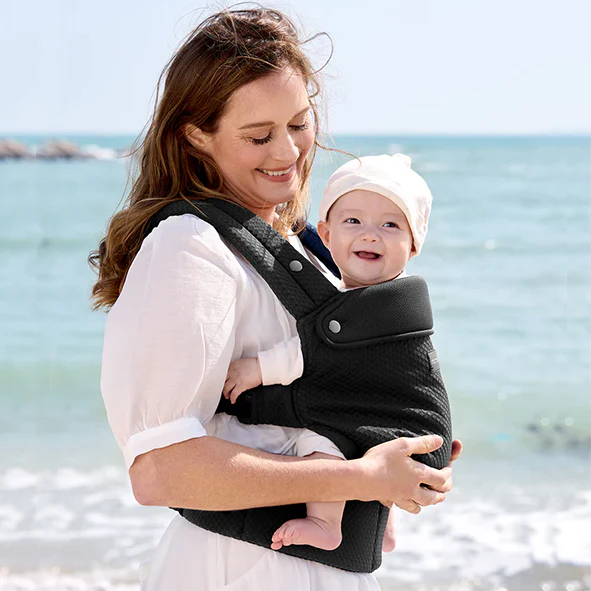 Una donna sorridente tiene un neonato in un marsupio scuro sulla spiaggia, vicino alla riva del mare.