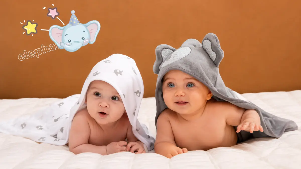 Dois beb&ecirc;s deitados em roupa de cama branca, cobertos com toalhas com capuz com motivo de elefante.