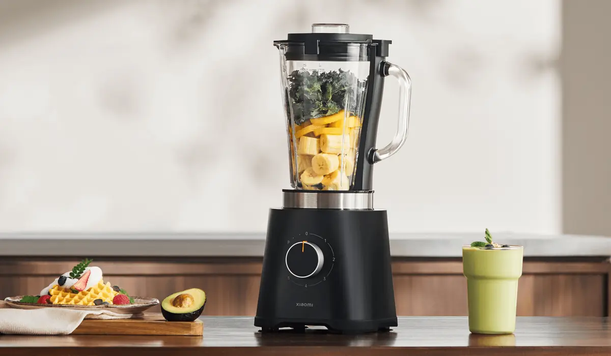 Blender Xiaomi z owocami (banan, mango, jarmuż) w kielichu, ustawiony na stole obok filiżanki napoju i awokado, z widocznym pokrętłem na podstawie
