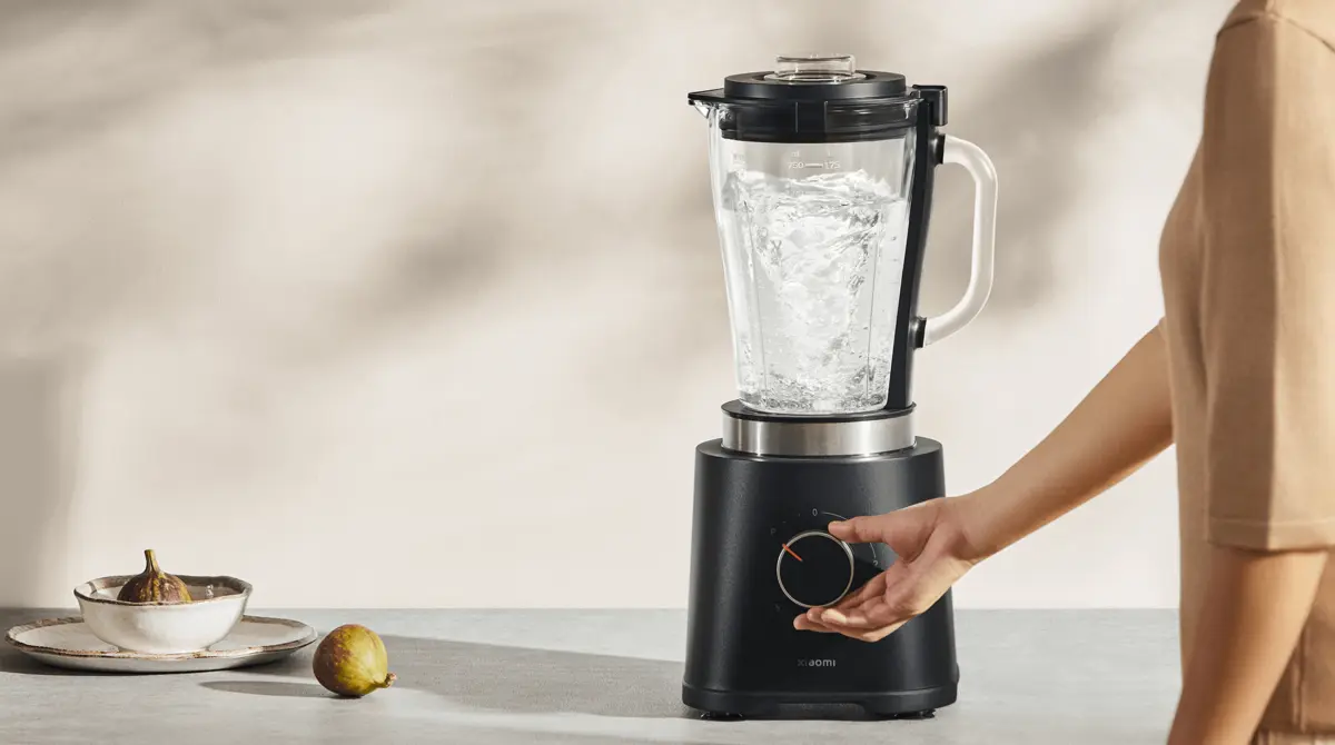 Blender Xiaomi z wodą w kielichu, ustawiony na stole, z ręką regulującą prędkość za pomocą pokrętła, obok figi na talerzu