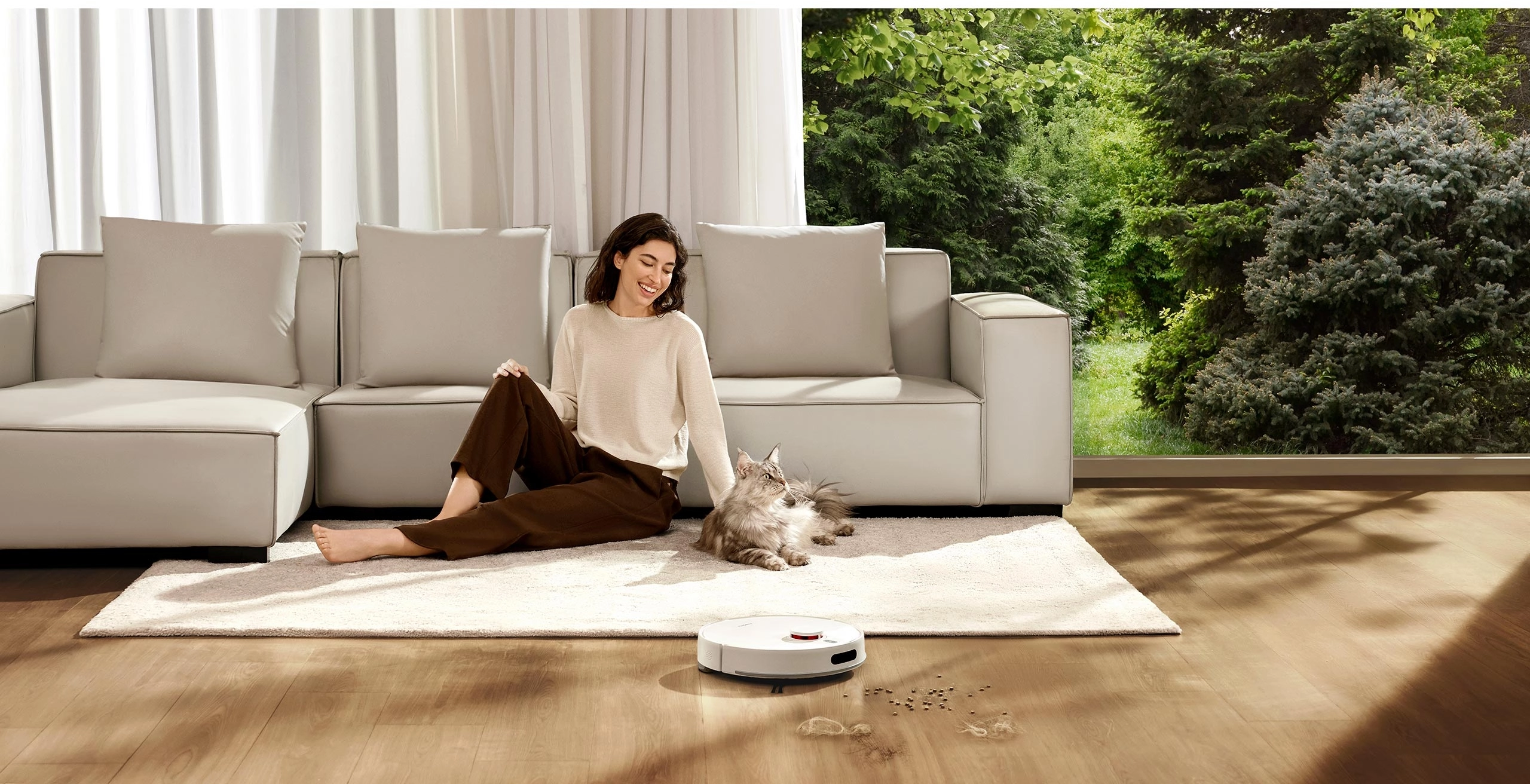 Робот Xiaomi Vacuum H40 пылесосит просторную гостиную, а женщина сидит на ковре, гладя кота, с видом на сад через окно