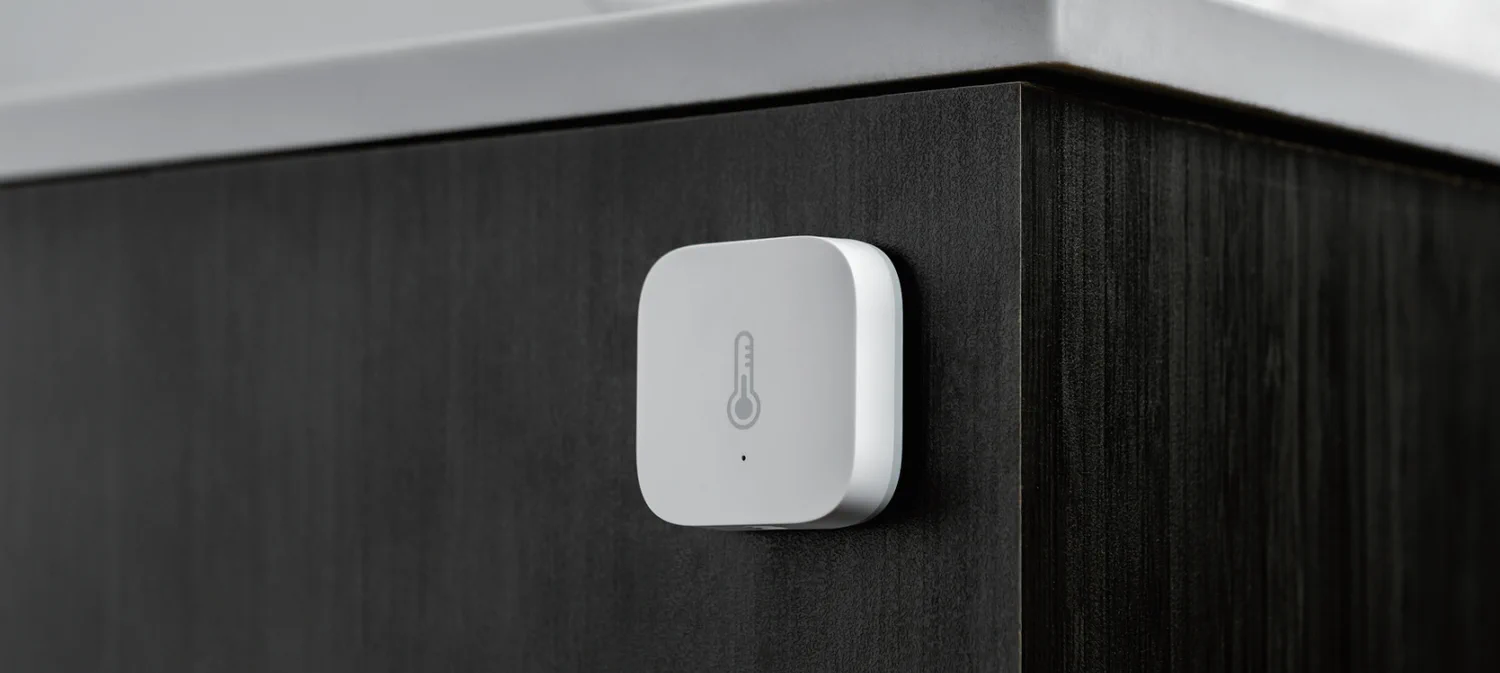 Czujnik temperatury Aqara zamontowany na ciemnej powierzchni szafki, monitorujący temperaturę