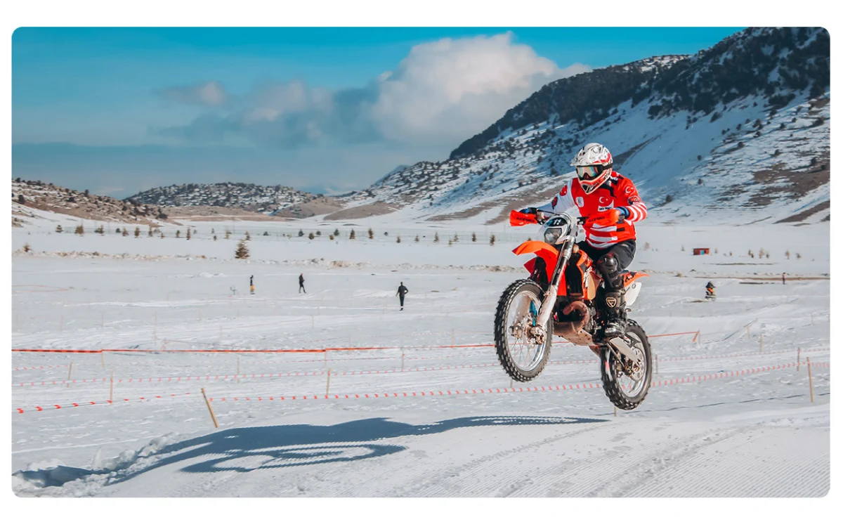 motocyklista w czerwonej koszulce, wykonujący skok na pomarańczowym motocyklu crossowym na śniegu, w tle góry