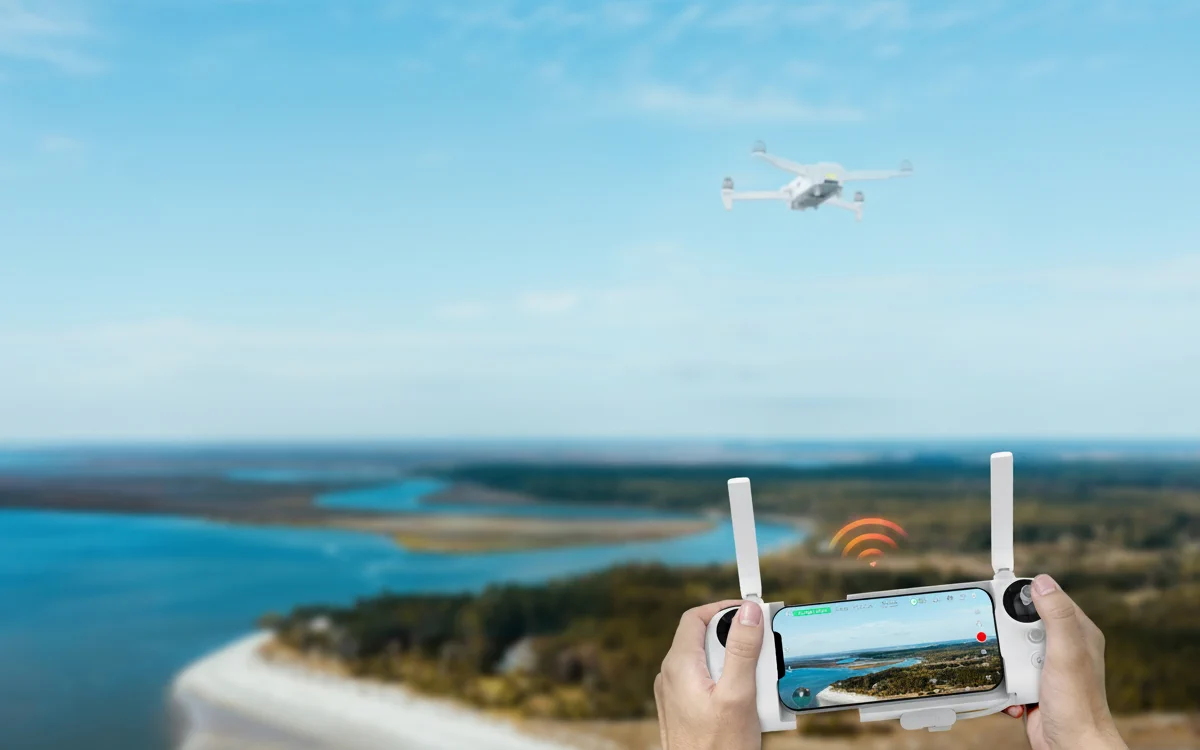 ręce sterujące dronem za pomocą kontrolera z podłączonym smartfonem, Dron FIMI X8 Tele w tle, leci nad wodą i zalesionym brzegiem