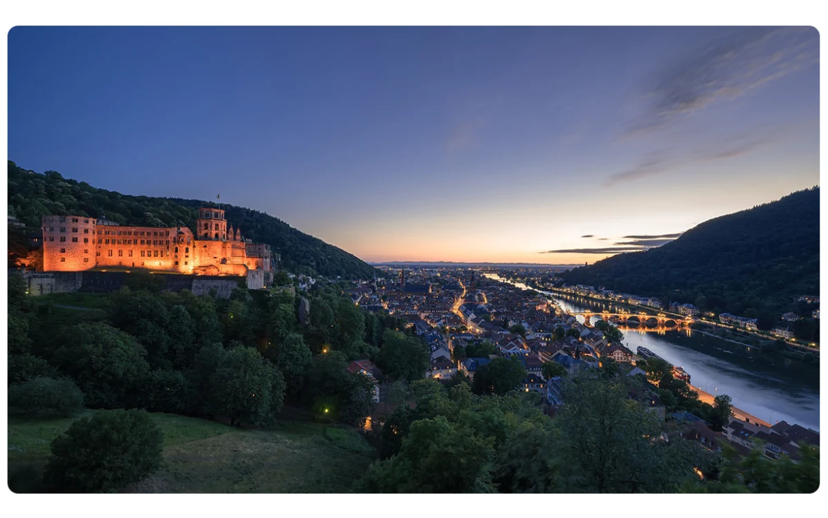 oświetlony zamek na zalesionym wzgórzu po lewej stronie oraz rozświetlone miasto leżące w dolinie rzeki