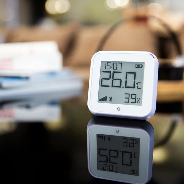 Termostat Shelly pokazujący 26°C i 39% wilgotności, ustawiony na lustrzanej powierzchni, tło lekko rozmyte
