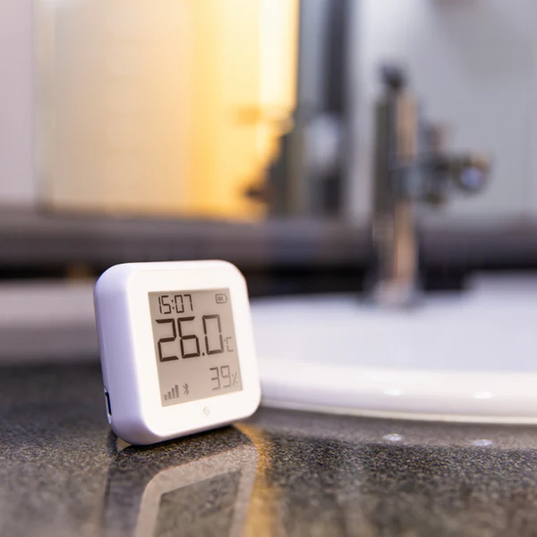 Termostat Shelly na blacie łazienkowym, pokazujący 26°C i 39% wilgotności, w nowoczesnej łazience