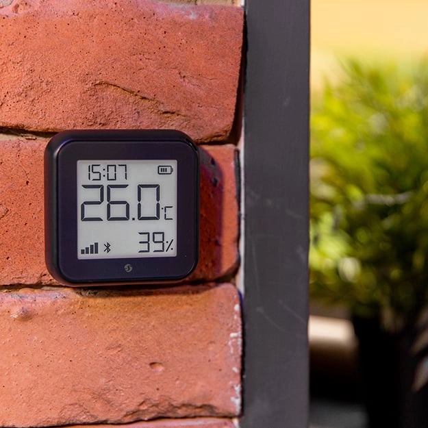Czarny czujnik temperatury i wilgotności zamocowany na ceglanej ścianie, z wyświetlaczem wskazującym 26°C i 39%