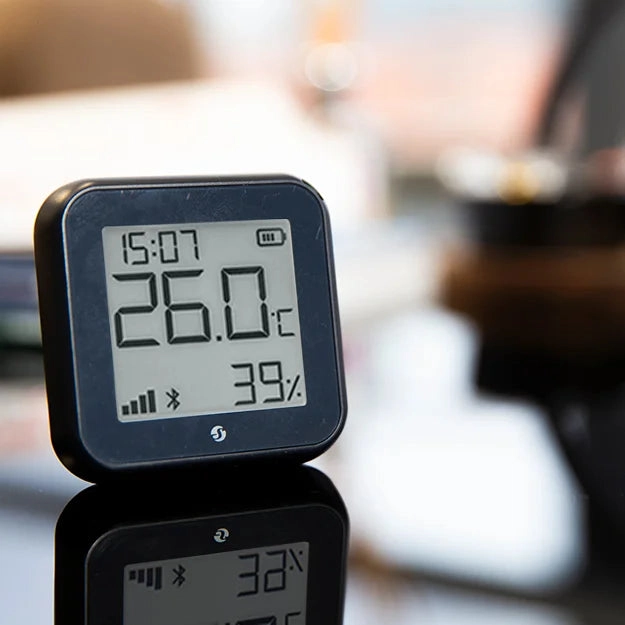 Czarny czujnik stojący na szklanej powierzchni, z wyświetlaczem pokazującym 26°C i 39% wilgotności