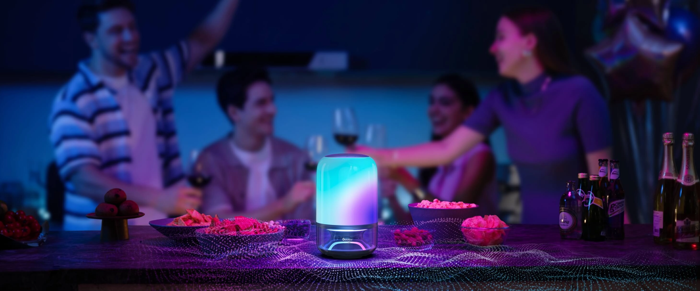 Lampa LED Govee H6020 Table Lamp 2 Pro świecąca niebiesko i różowo na stole z przekąskami podczas imprezy, w tle grupa osób rozmawiających w przyciemnionym pomieszczeniu