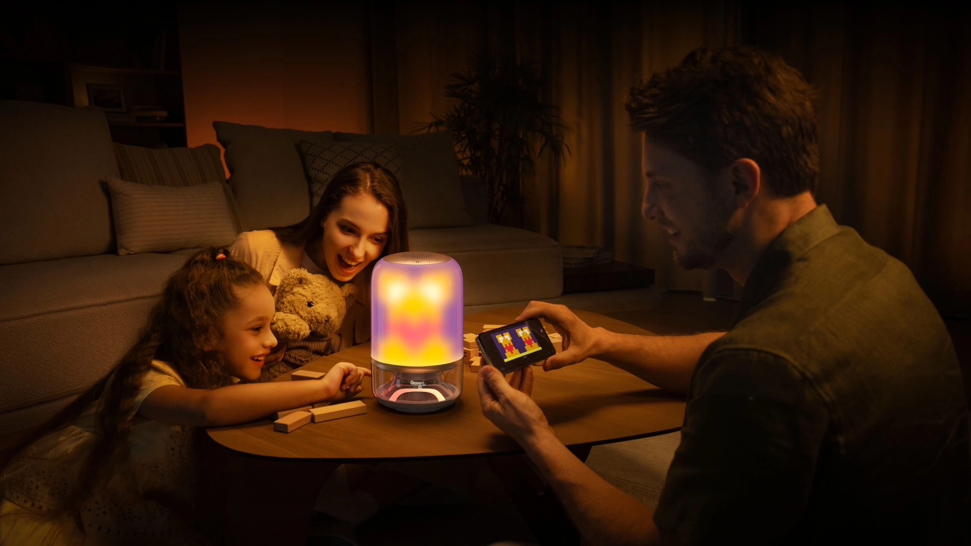 Rodzina z dzieckiem siedząca przy stole w ciepło oświetlonym salonie, dziecko bawi się przy lampie Govee H6020 świecącej żółto-fioletowym światłem, obok mężczyzna trzyma smartfon