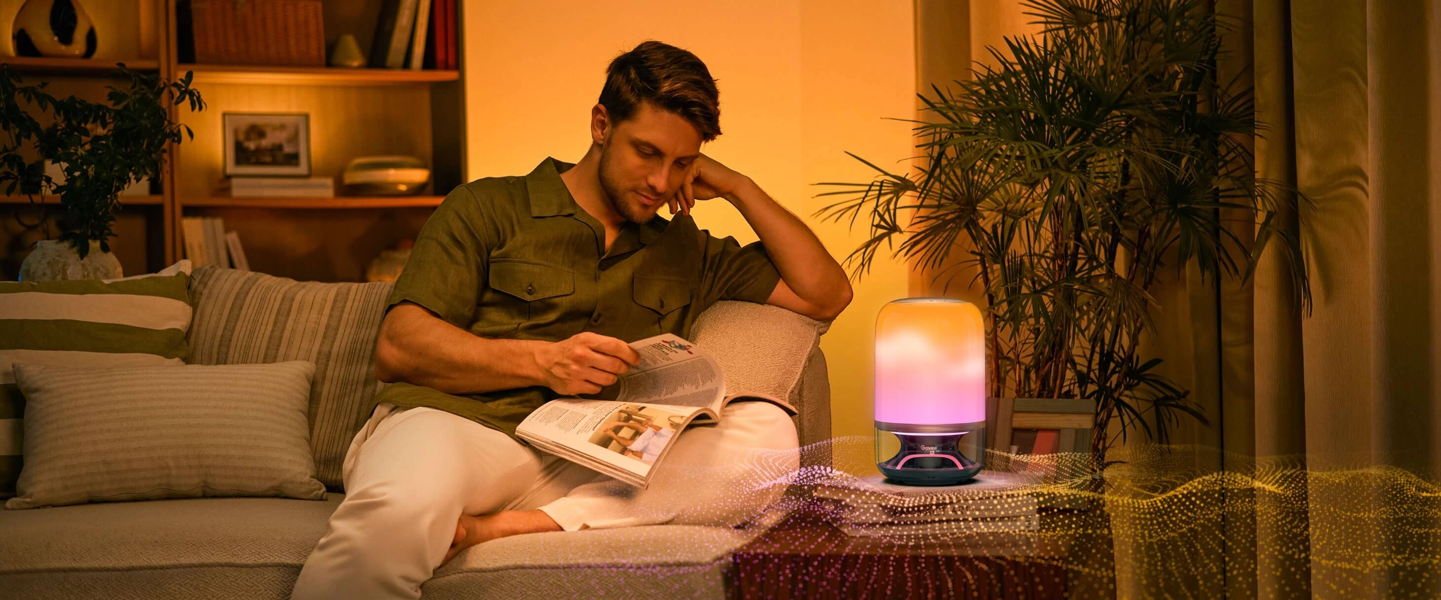 Mężczyzna siedzący na sofie w ciepło oświetlonym salonie czyta magazyn, obok na stoliku świeci lampa LED Govee H6020 w barwach żółto-różowych