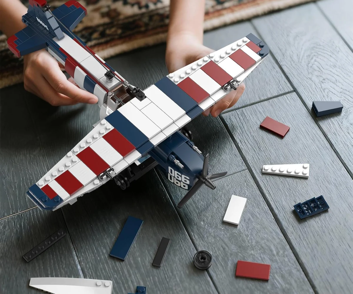 M&atilde;os de uma crian&ccedil;a montando um modelo de avi&atilde;o feito de blocos no ch&atilde;o, rodeadas por pe&ccedil;as soltas da constru&ccedil;&atilde;o