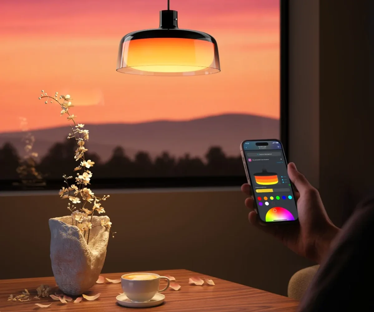 A hand holding a smartphone with an app for controlling the colour of a Govee H60C1 pendant lamp against a sunset background