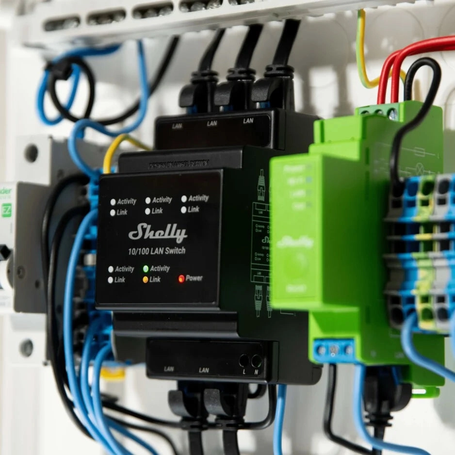 Wnętrze szafki elektrycznej z przełącznikiem LAN Shelly, widoczna obudowa z niebieskimi i zielonymi przewodami