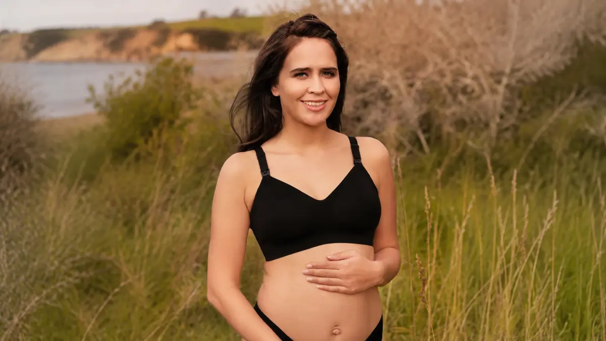Mulher com sutiã de gravidez preto está ao ar livre, com a mão a envolver a barriga, com relva e um lago ao fundo.