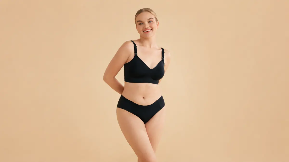 Mulher com sutiã de gravidez preto está sobre um fundo bege, com as mãos atrás das costas, a sorrir, com a silhueta completa visível.
