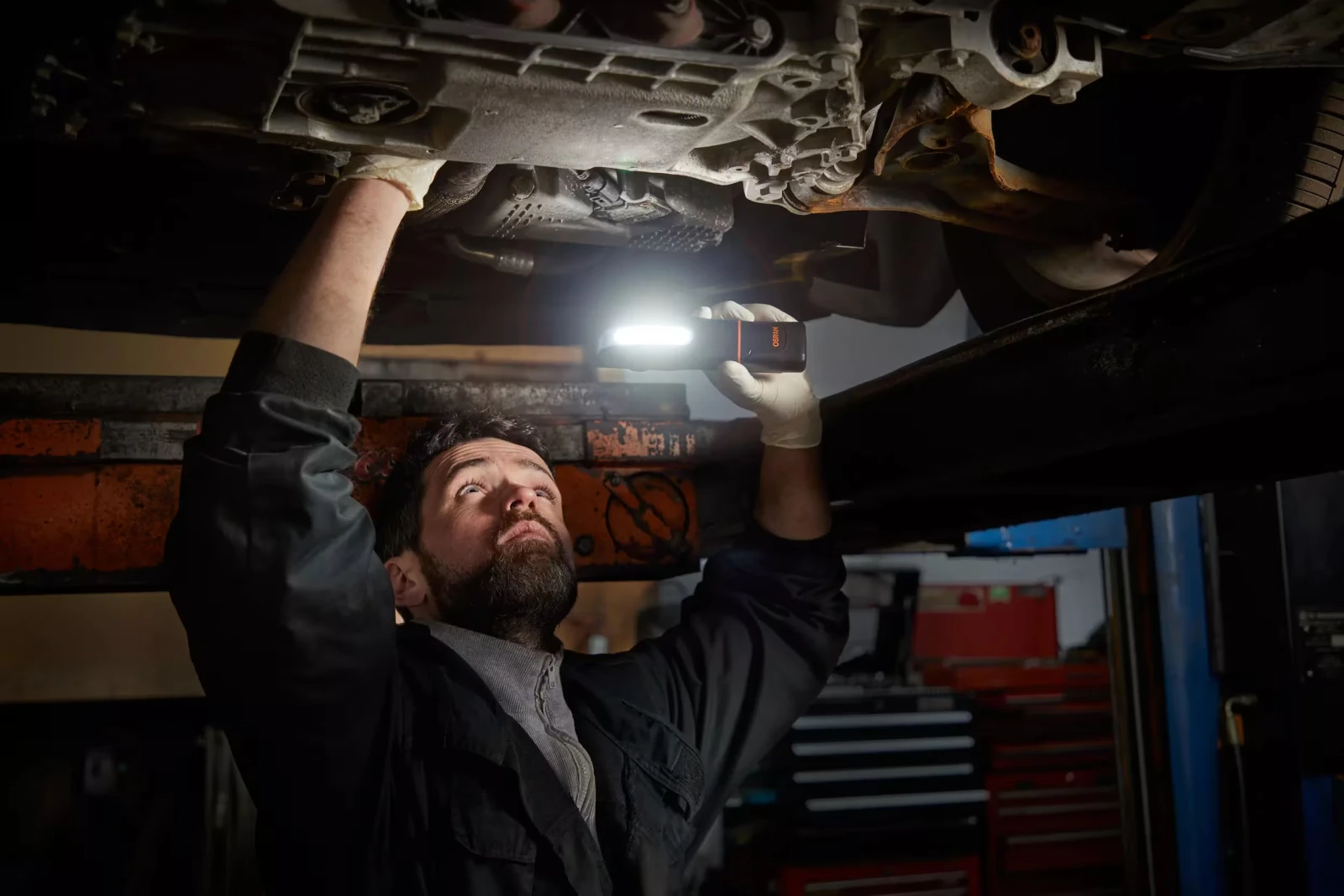 Mechanik w warsztacie oświetla podwozie samochodu na podnośniku za pomocą ręcznej lampy warsztatowej LED