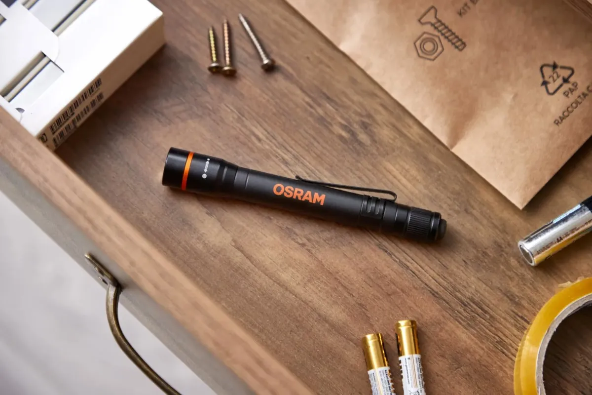 Czarna latarka OSRAM z pomarańczowym pierścieniem leżąca na drewnianym blacie obok baterii AAA i śrub.