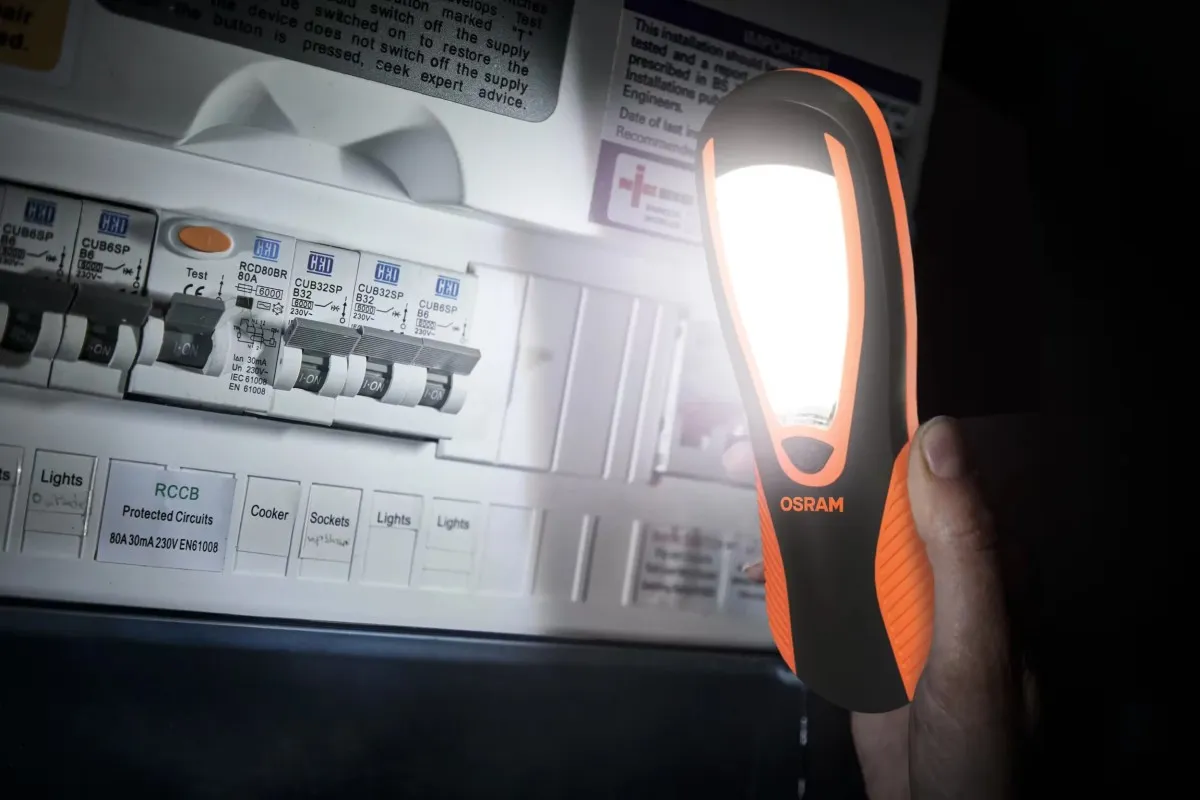 Lampa warsztatowa OSRAM oświetlająca białą rozdzielnię elektryczną trzymana w dłoni w ciemnym pomieszczeniu