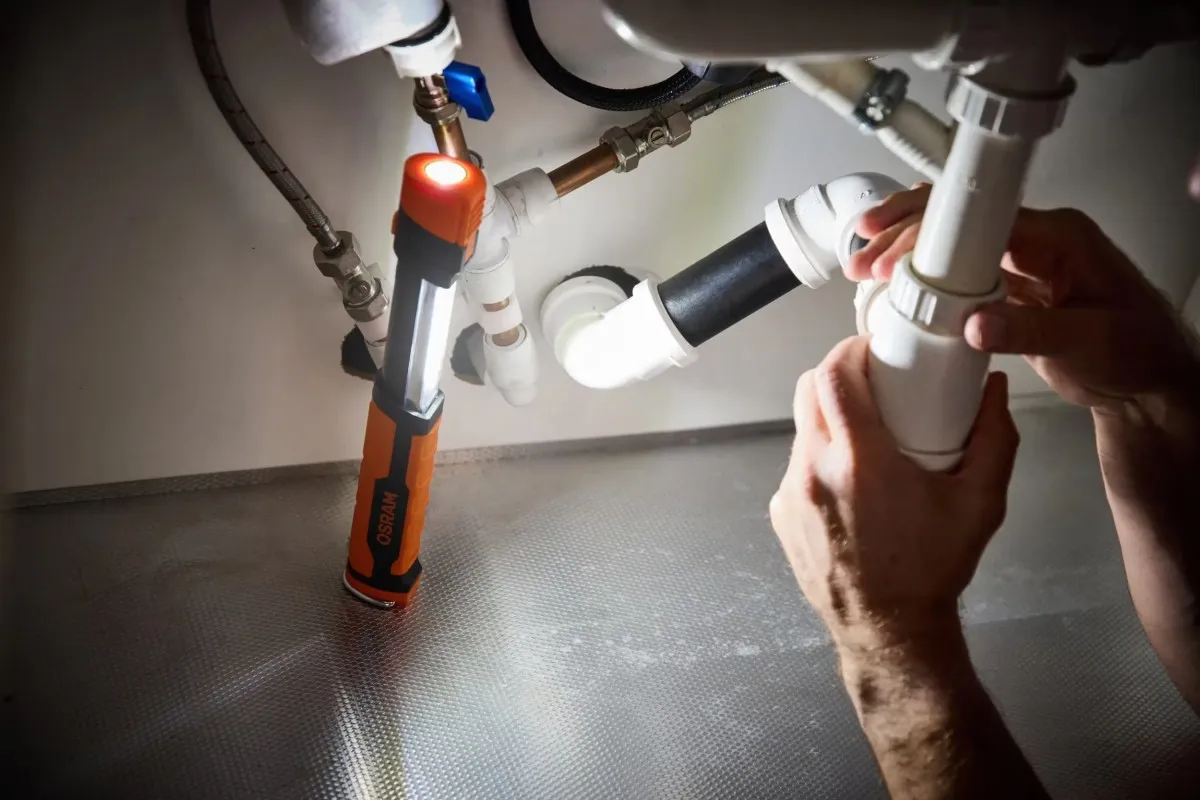 Lampa OSRAM przytwierdzona magnesem pod zlewem, oświetlająca rury podczas naprawy hydraulicznej.
