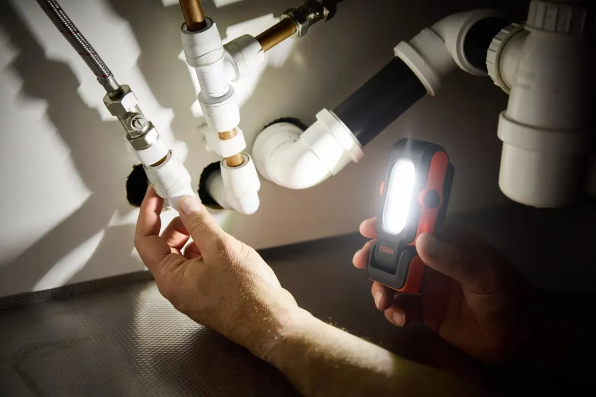Osoba doświetla lampą OSRAM instalację wodną pod zlewem podczas prac naprawczych.