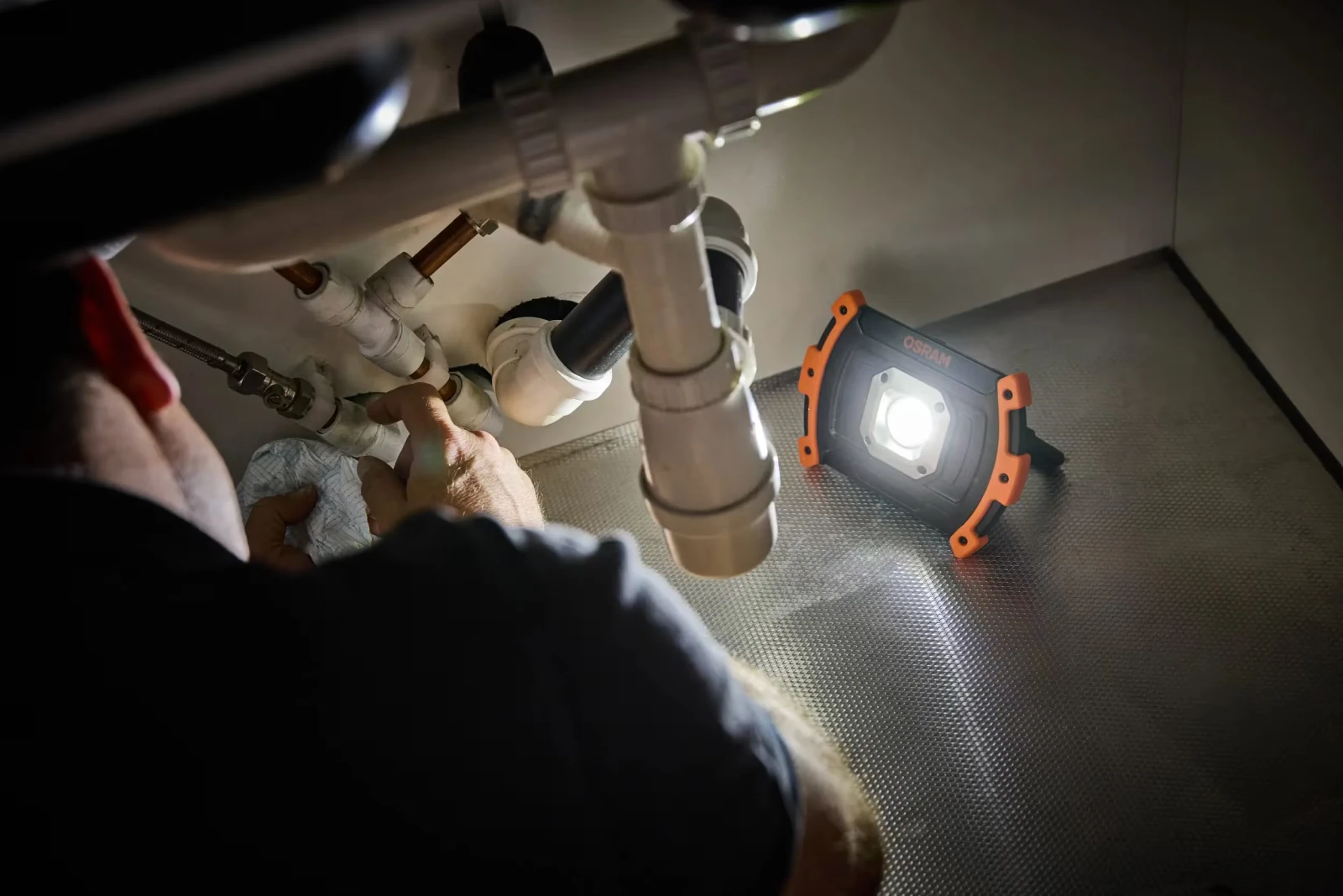 Lampa warsztatowa LED stoi pod zlewem, oświetlając rury odpływowe podczas prac hydraulicznych