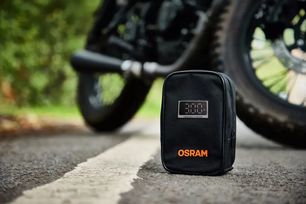 Czarny pokrowiec z logo OSRAM i wyświetlaczem cyfrowym stojący na asfalcie obok koła motocykla.