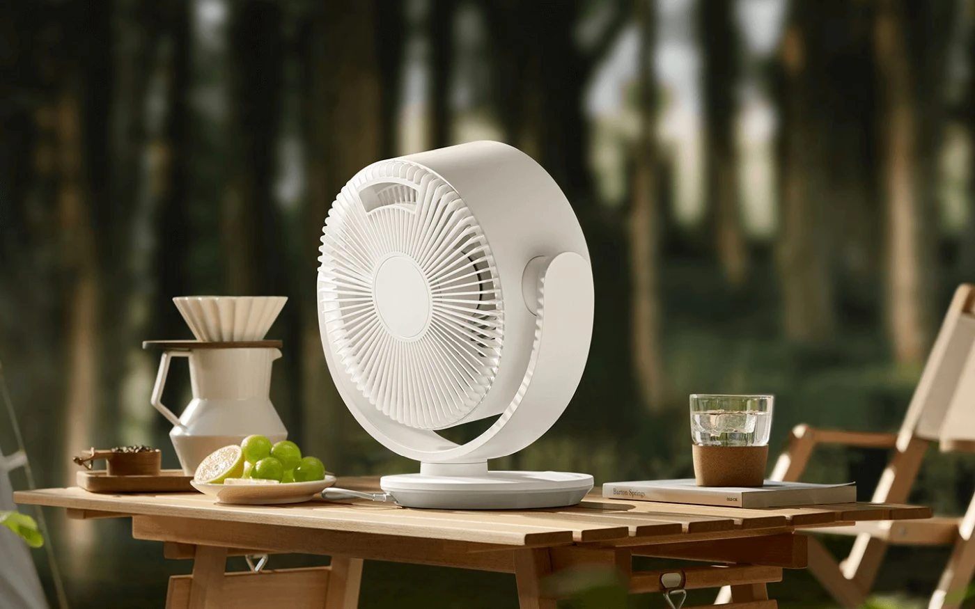 Ventilador Xiaomi blanco sobre una mesa de madera de camping con un bosque de fondo, junto a una jarra de café y un plato con fruta