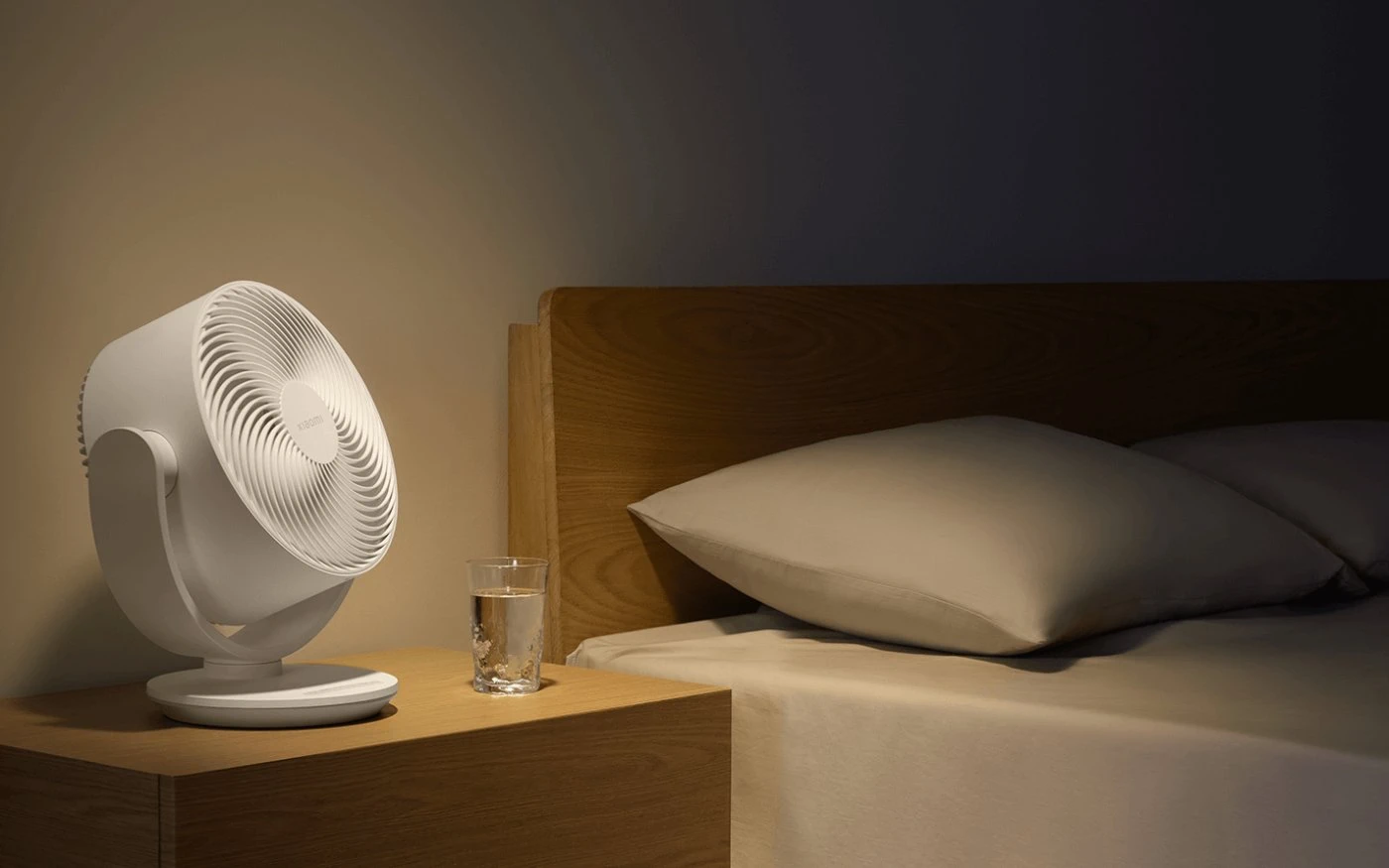 Ventilador Xiaomi blanco sobre una mesita de noche de madera junto a un vaso de agua en un dormitorio acogedor y bien iluminado