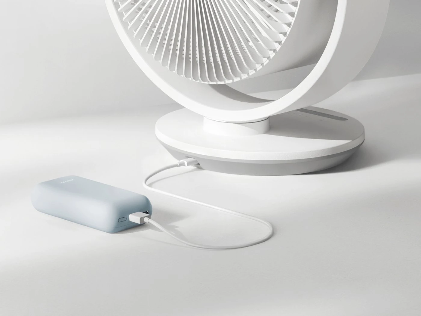 Ventilador blanco conectado con un cable USB a una powerbank azul claro sobre un fondo claro