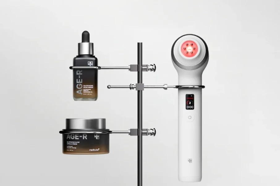 Dispositivo branco com cabeça circular e dois frascos cosméticos pretos sobre um suporte metálico.