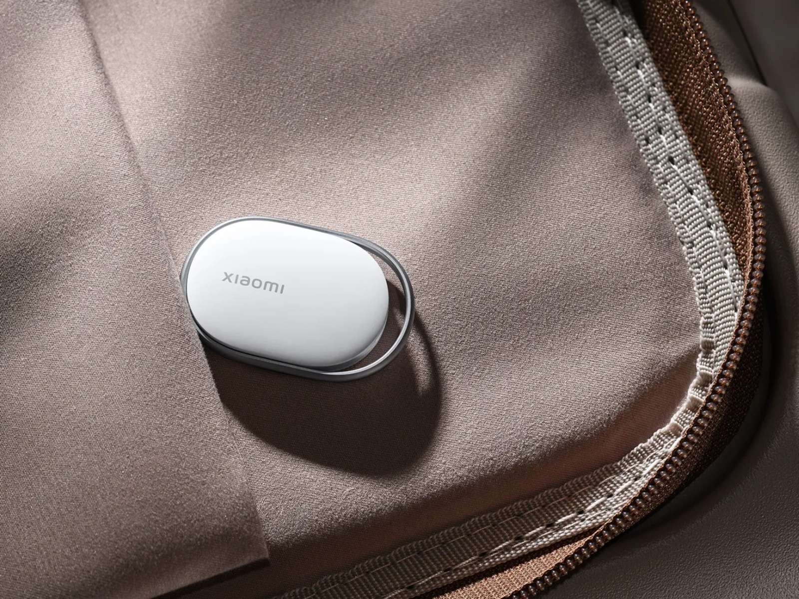 Белый овальный локатор Xiaomi с металлической рамкой, лежащий внутри коричневой дорожной сумки