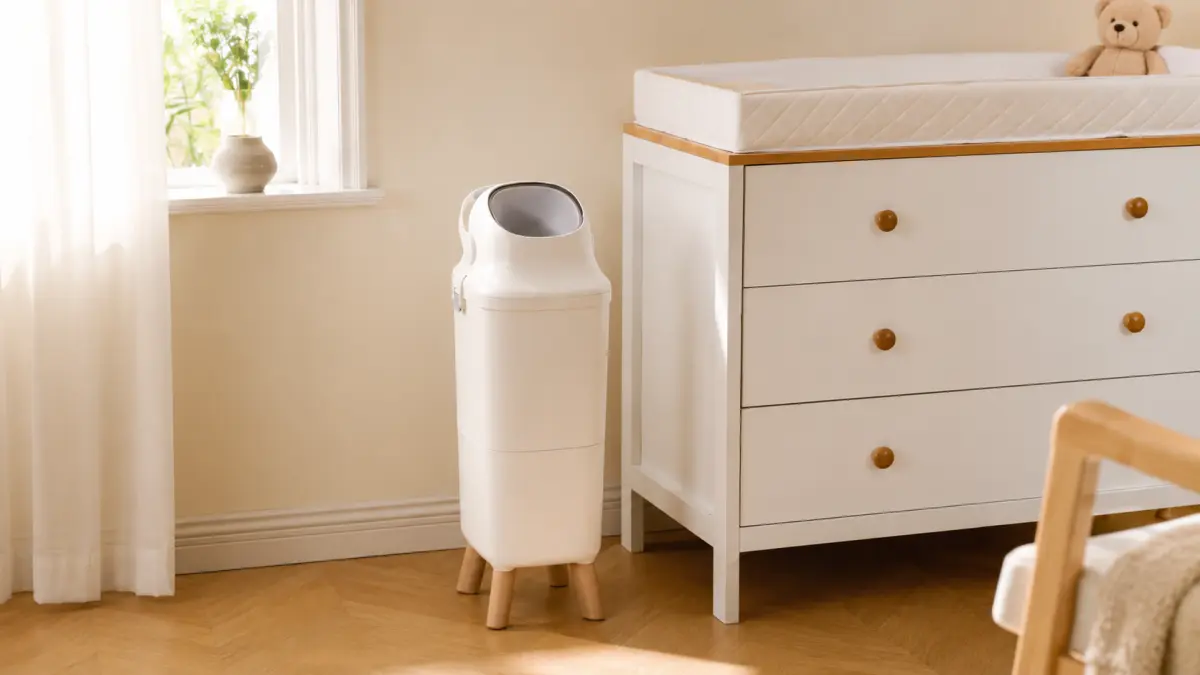 Um cesto branco para fraldas est&aacute; ao lado de uma c&oacute;moda com trocador num quarto infantil claro.