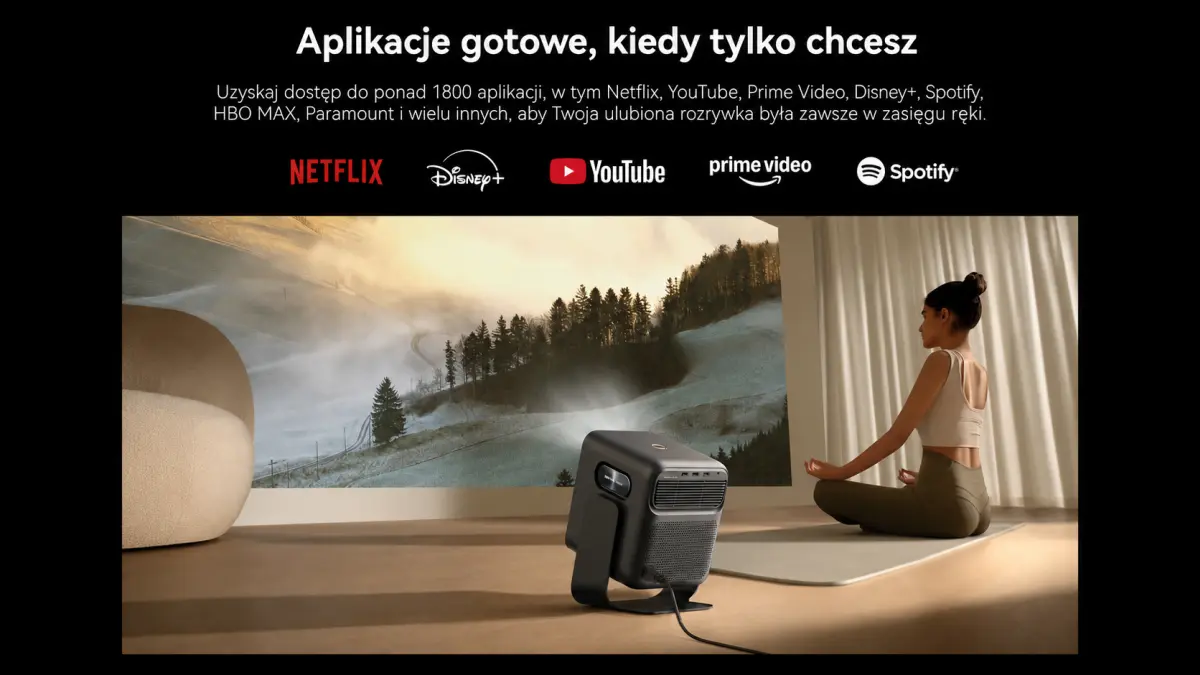 Projektor Mozart 1S wyświetlający górski krajobraz w pomieszczeniu, obok kobiety siedzącej na macie.
